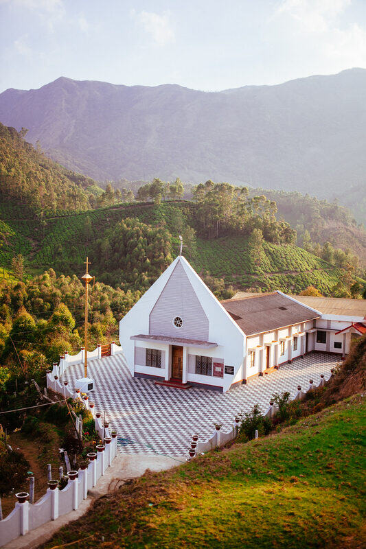 A white Catholic chapel sits below tea-covered mountains near Munnar Top Station, reflecting the Catholic and Portuguese layers in the region.