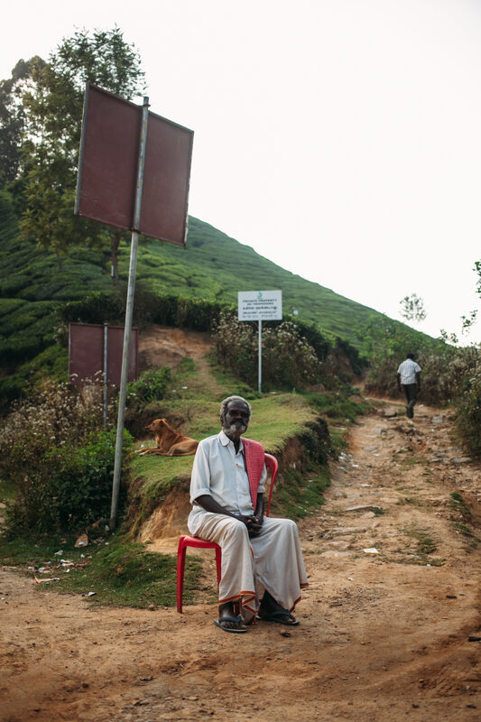 A blind man sits quietly on a red chair beside a Munnar hill road, listening to the world around him.