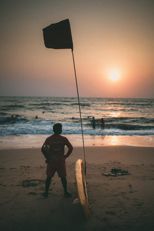 A Goa beach lifeguard board and safety flag turn into a sunset silhouette above the surf.