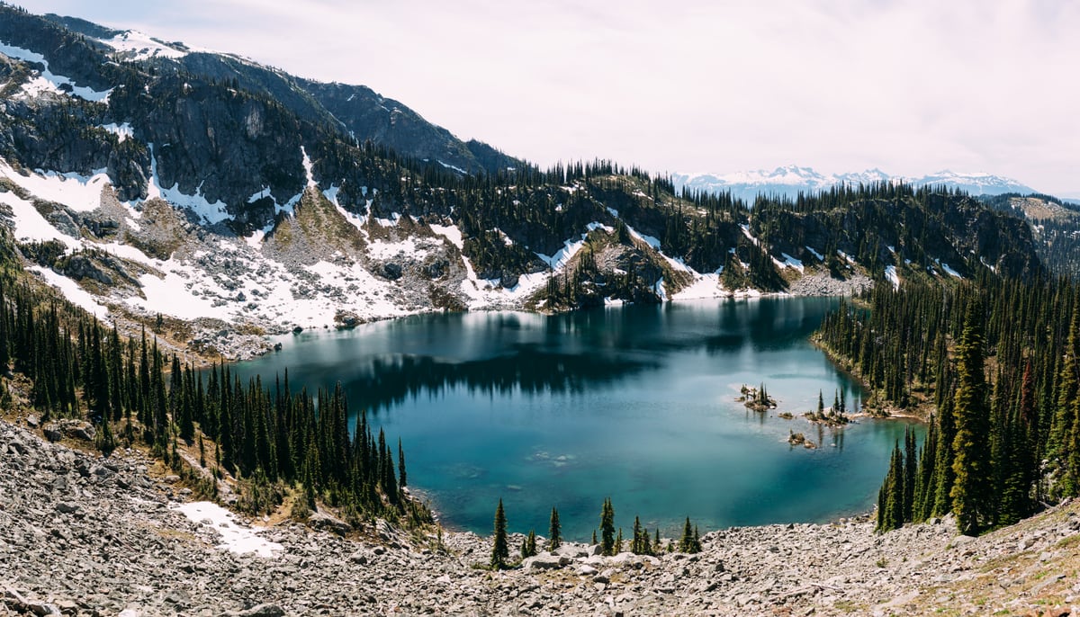 A wide Mount Revelstoke alpine lake held by rock, snow and summer sky.