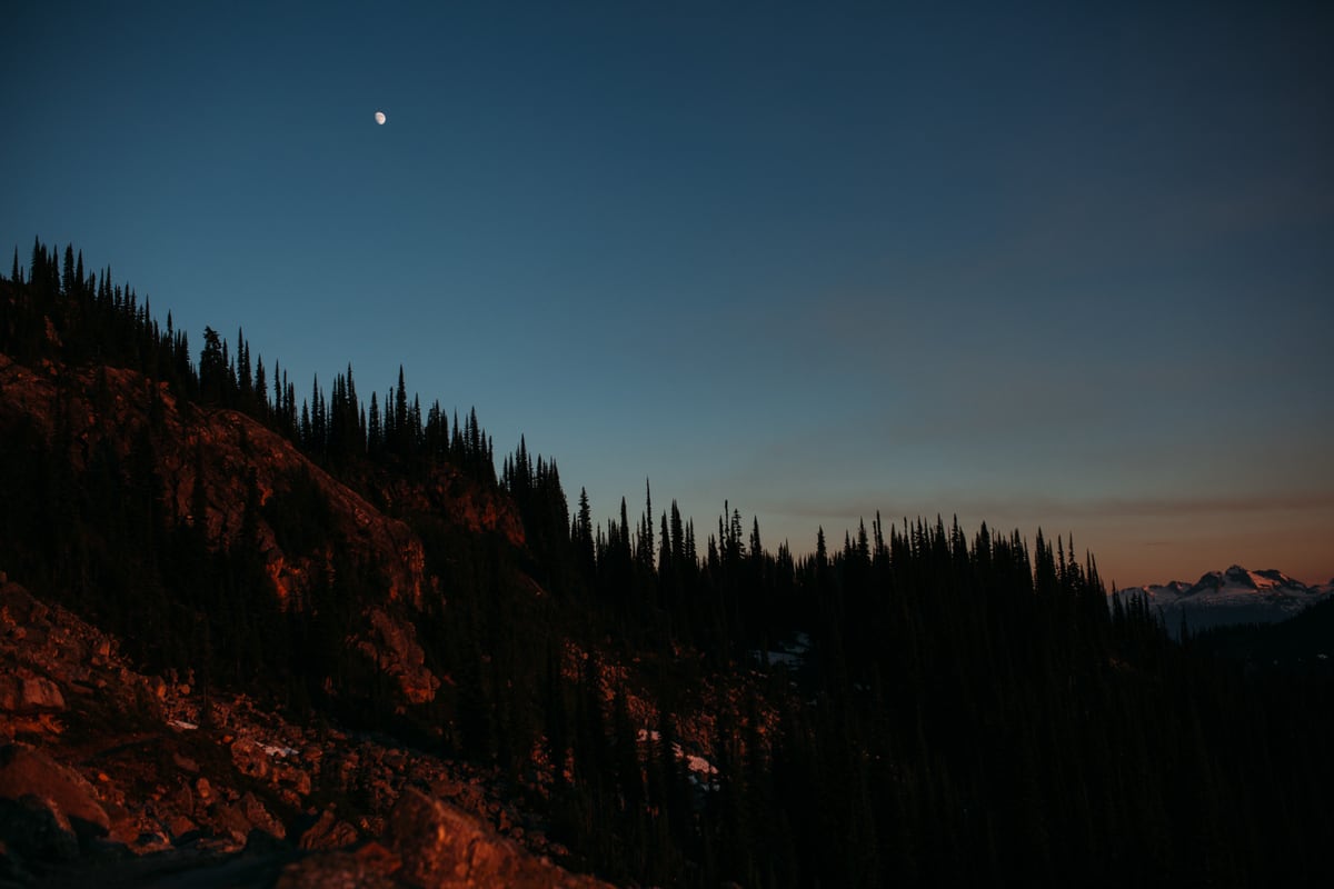 The moon rises above a dark Mount Revelstoke ridge at blue hour.