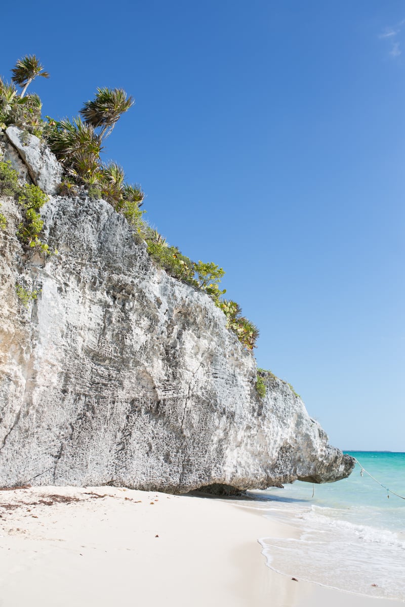 White Tulum limestone, blue water and cliff plants under clear sky.