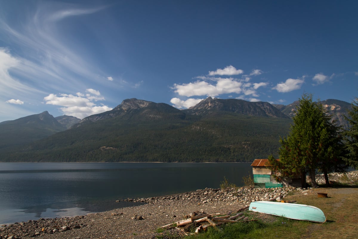 A green canoe rests on a rocky shore below mountain water and sky in the Kootenays near Kaslo, BC.