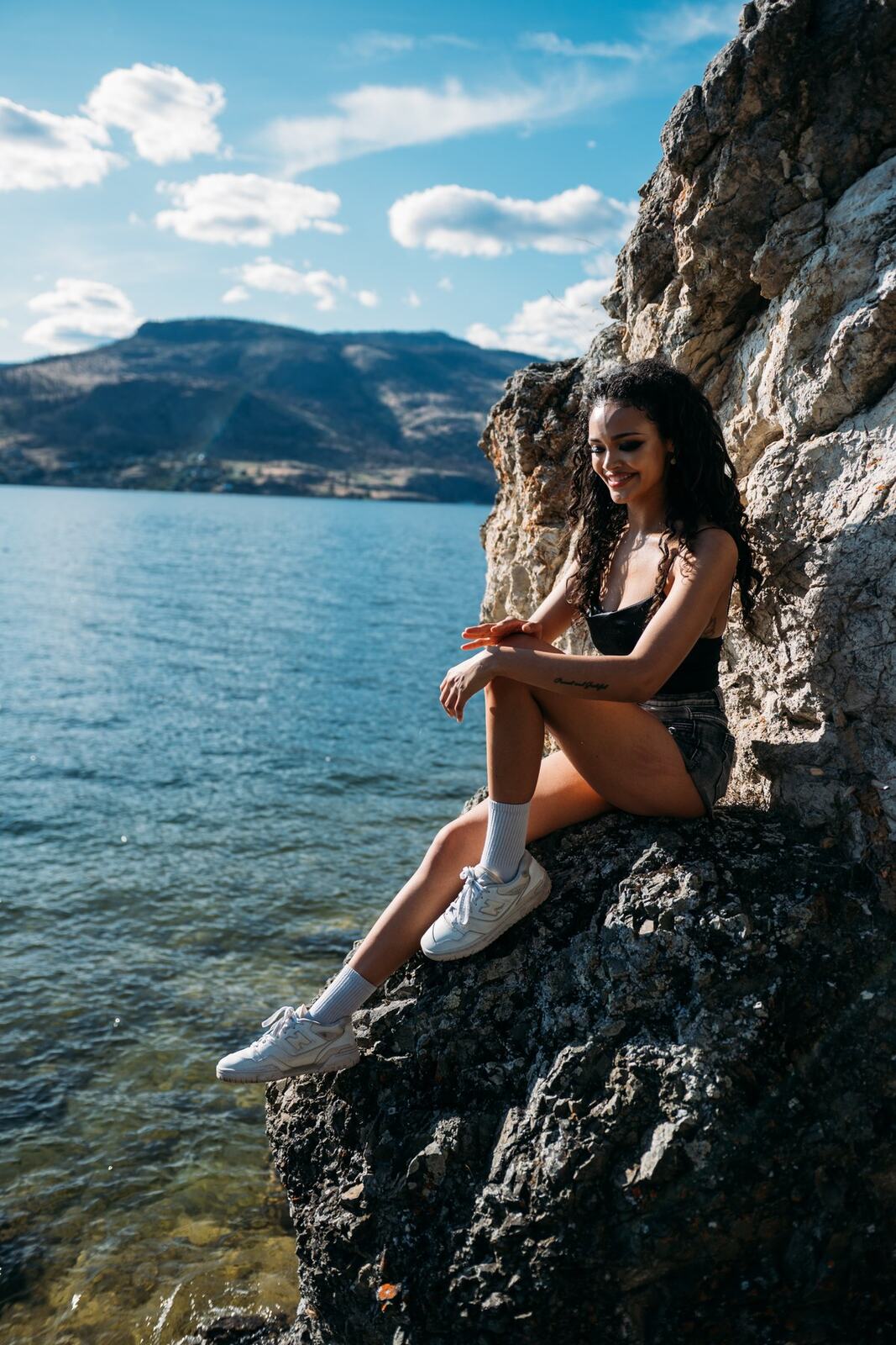 Alex by the Lake portraits story frame 3: Okanagan Lake portraits in Kelowna with black styling, rock, water, wind, direct expression and a cinematic beauty-portrait edge.